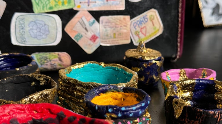 A collection of brightly painted and gold leafed pots photographed in from of a suitcase covered in hand-drawn luggage labels